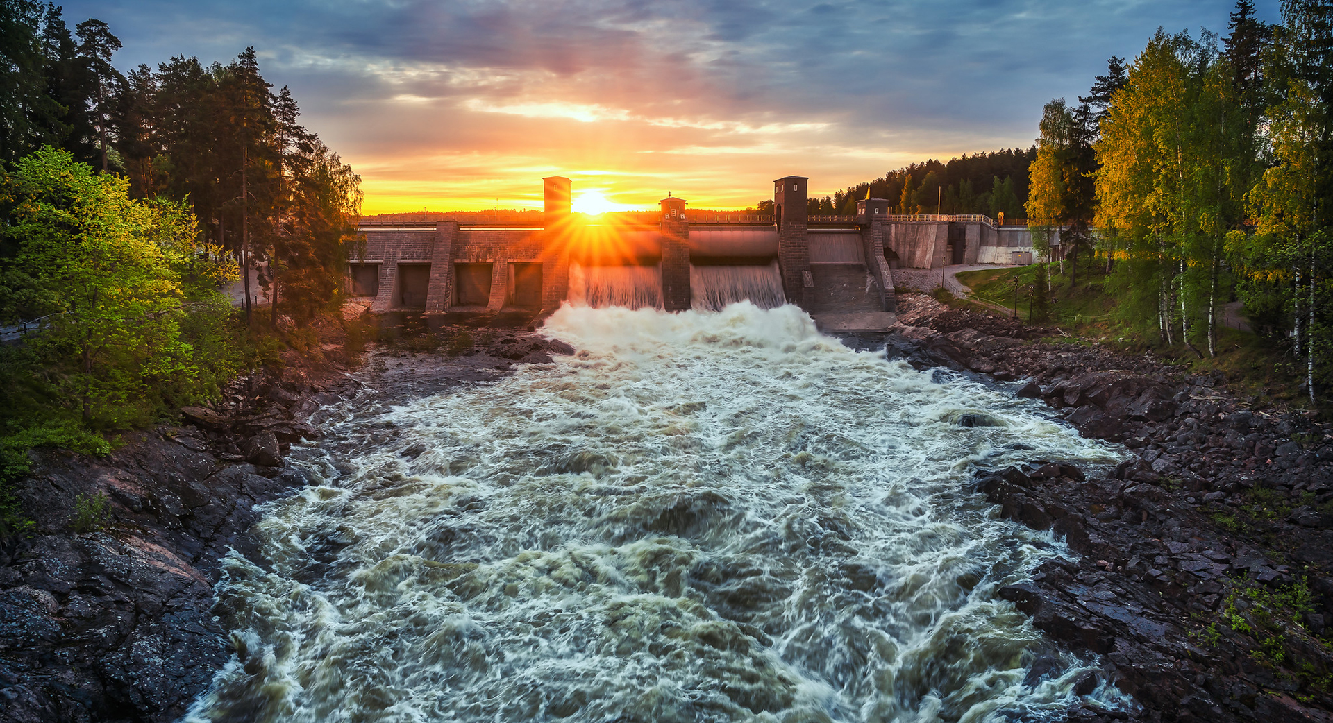 The rapids of Imatrankoski in full surge, with forest along the banks. In the background, a dam with the sun setting behind it.